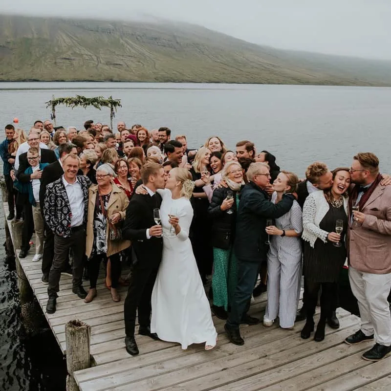 Brúðhjón og gestir fagna á trébryggju við fjörðinn.
